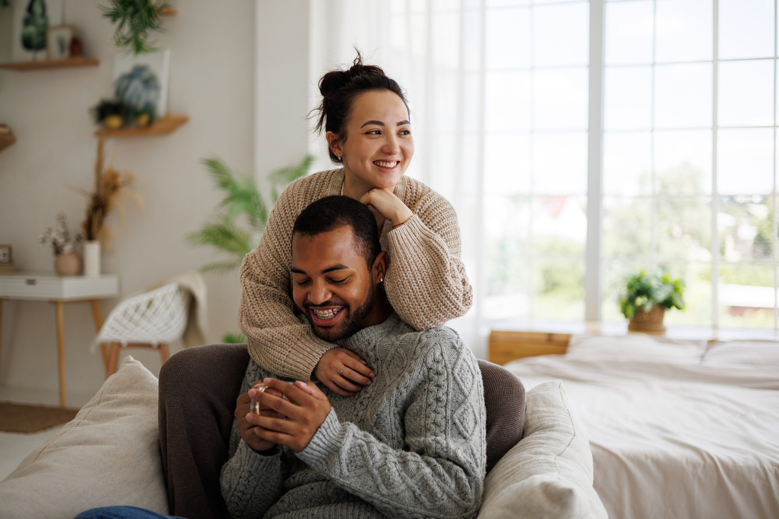 Smiling young asian woman in sweater hugging african american boyfriend holding coffee at home with proper furnace care